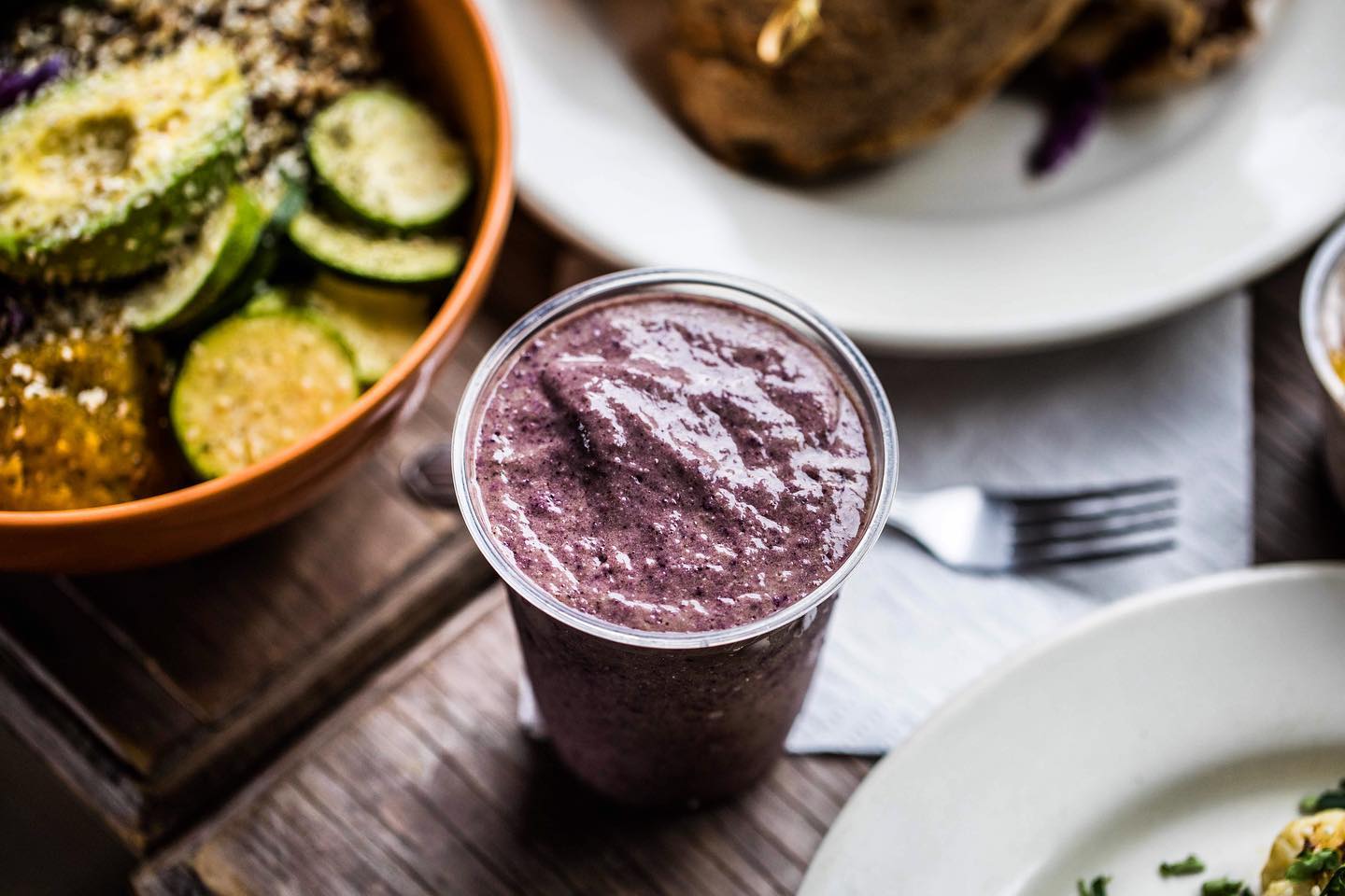 Purple smoothie in a clear cup on a wooden table with plates of food in the background
