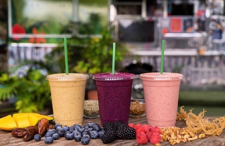 Three smoothie cups with straws on a wooden surface with fruits and vegetables.
